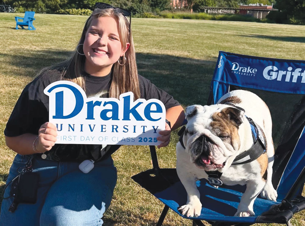 A smiling student holds a Drake University sign while sitting on the grass next to an English bulldog.