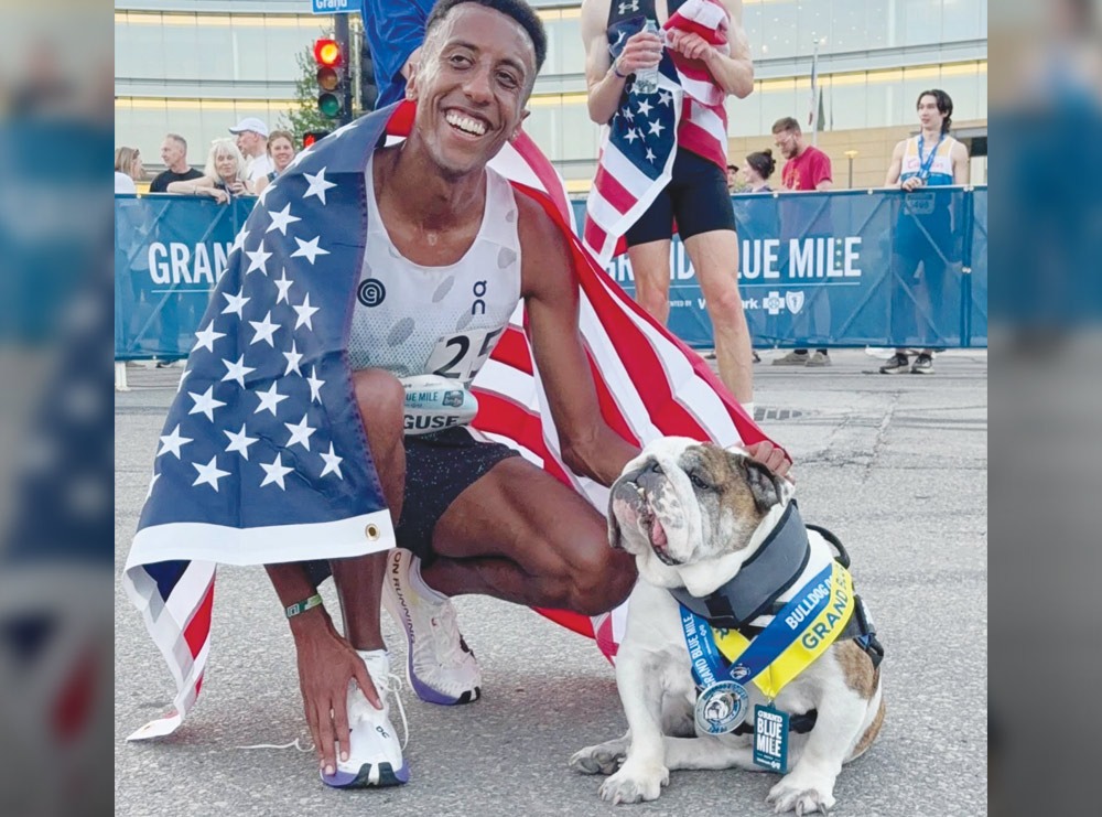 A runner draped in an American flag kneels to pose with an English bulldog wearing a medal and sash.