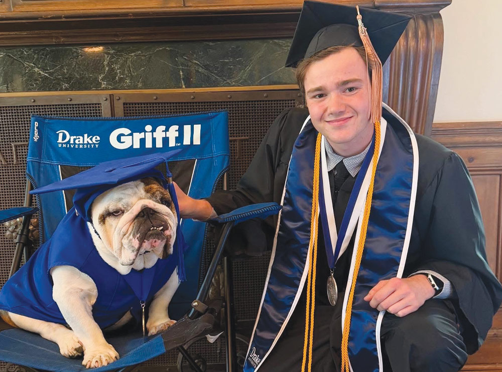 A graduate in cap and gown poses indoors next to an English bulldog wearing matching graduation attire.