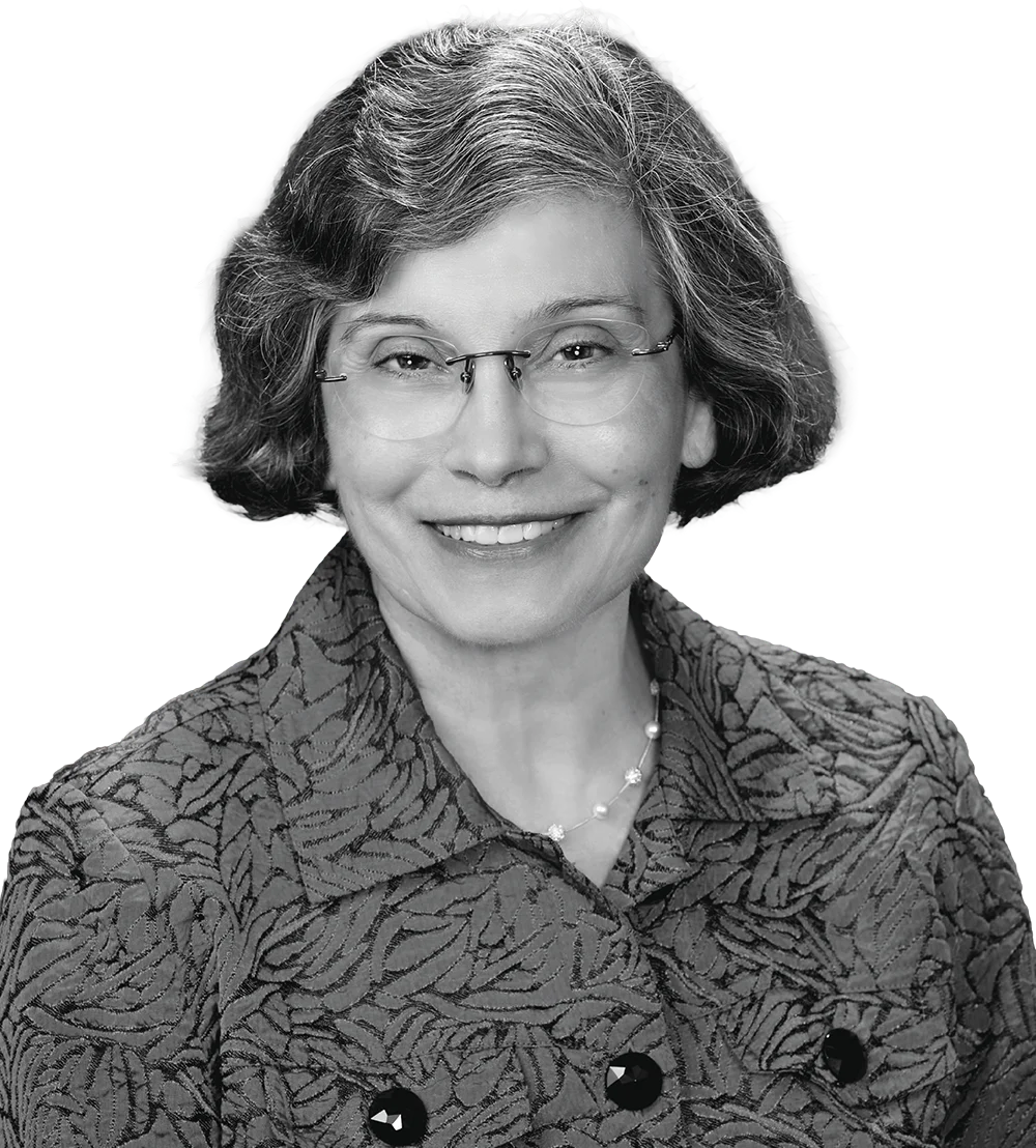 Black-and-white studio portrait of a smiling Barbara Quijano Decker with short hair and glasses wearing a patterned jacket.
