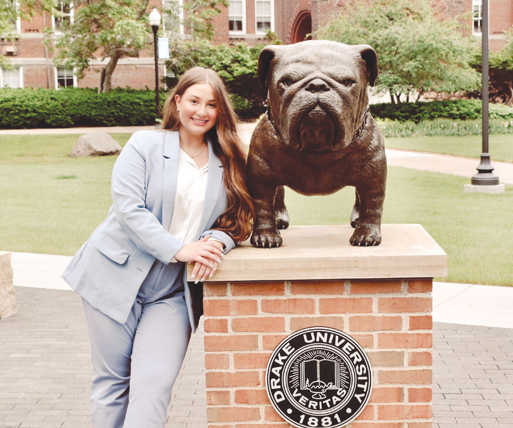 Samra Aksamovic in a light blue suit poses leaning against a brick pedestal supporting a bronze bulldog statue.