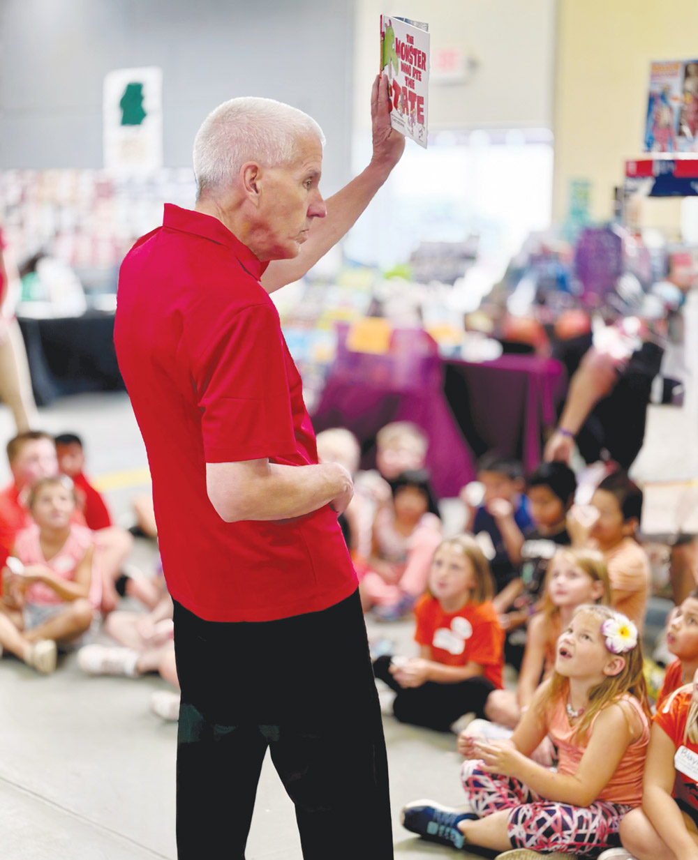 Joe Graves in a red shirt holds up a children's book while reading to a group of young students seated on the floor.