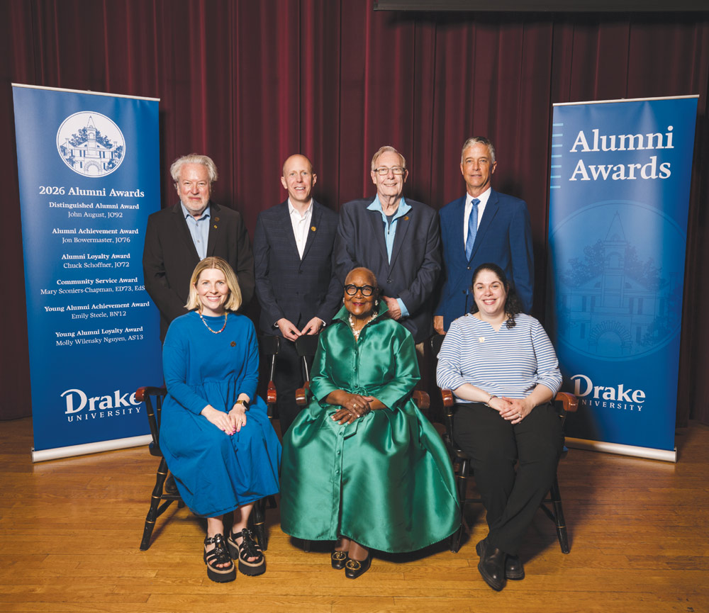 Seven alumni pose together on stage during an awards ceremony in front of Drake Alumni Awards banners.