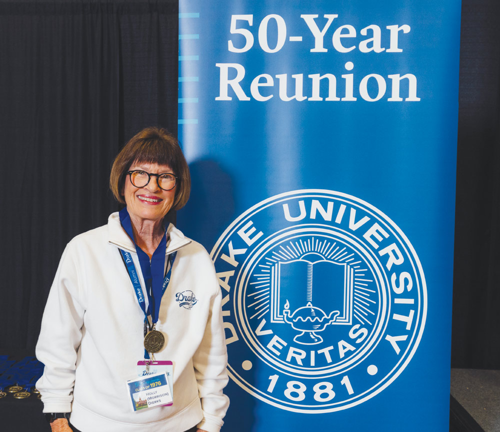 A smiling woman wearing glasses and a lanyard stands next to a "50-Year Reunion" Drake banner.