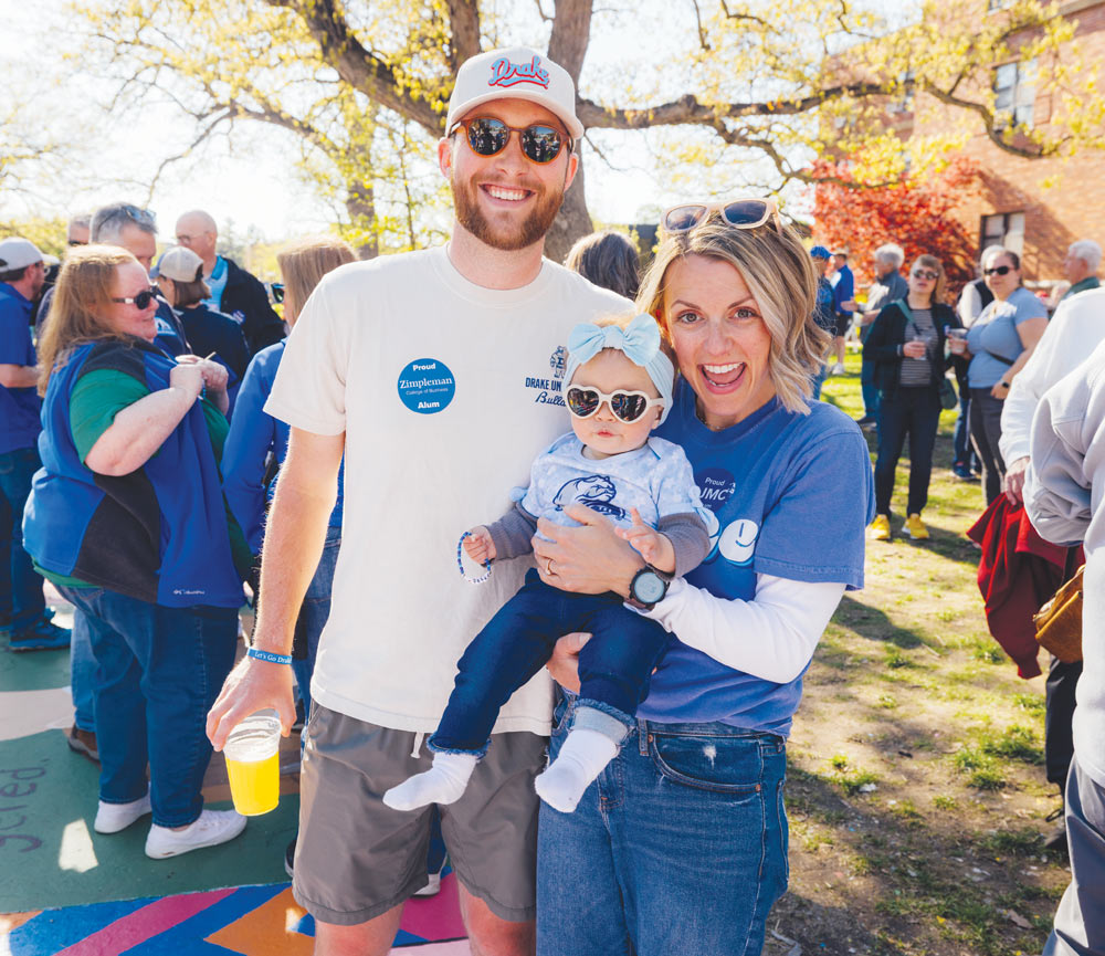 A smiling couple in sunglasses poses outdoors with their baby who is wearing heart-shaped sunglasses.