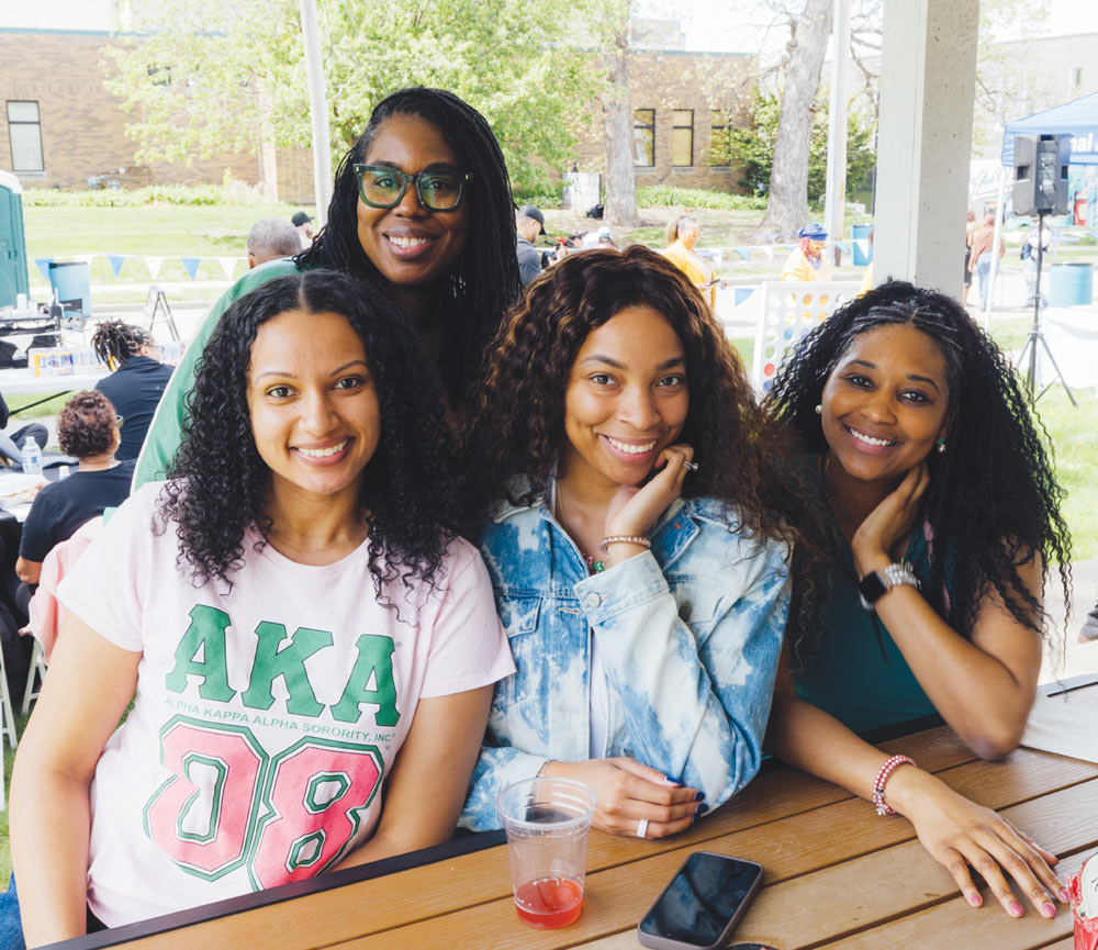 Four smiling women sitting together at an outdoor picnic table during a campus reunion event.
