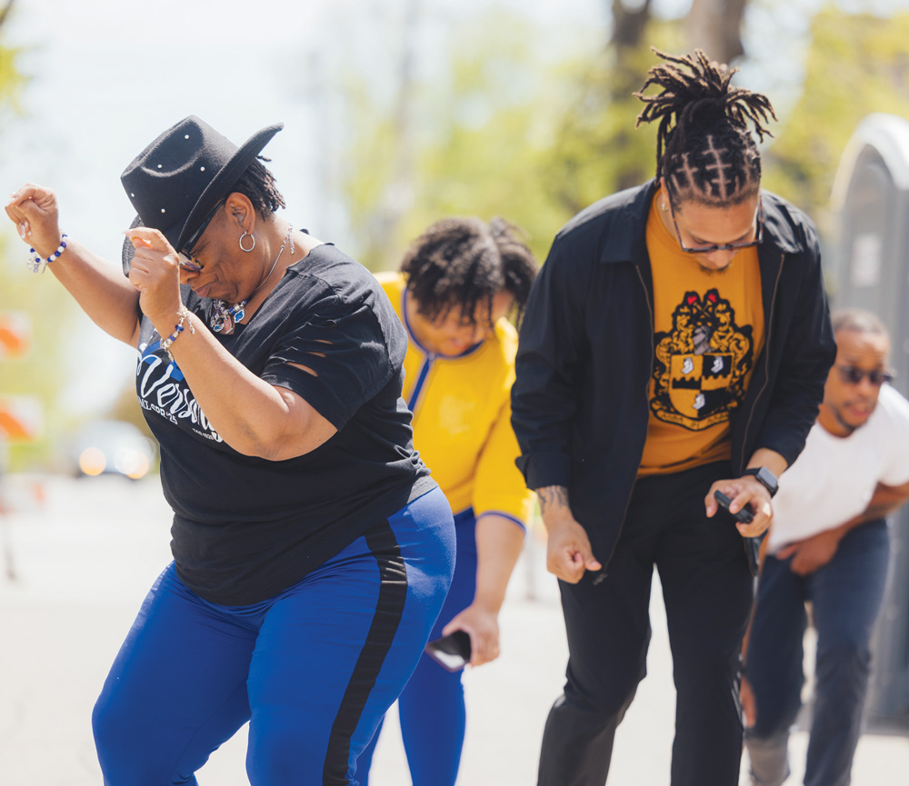 Four people dance outdoors together on a paved walkway during a campus event.