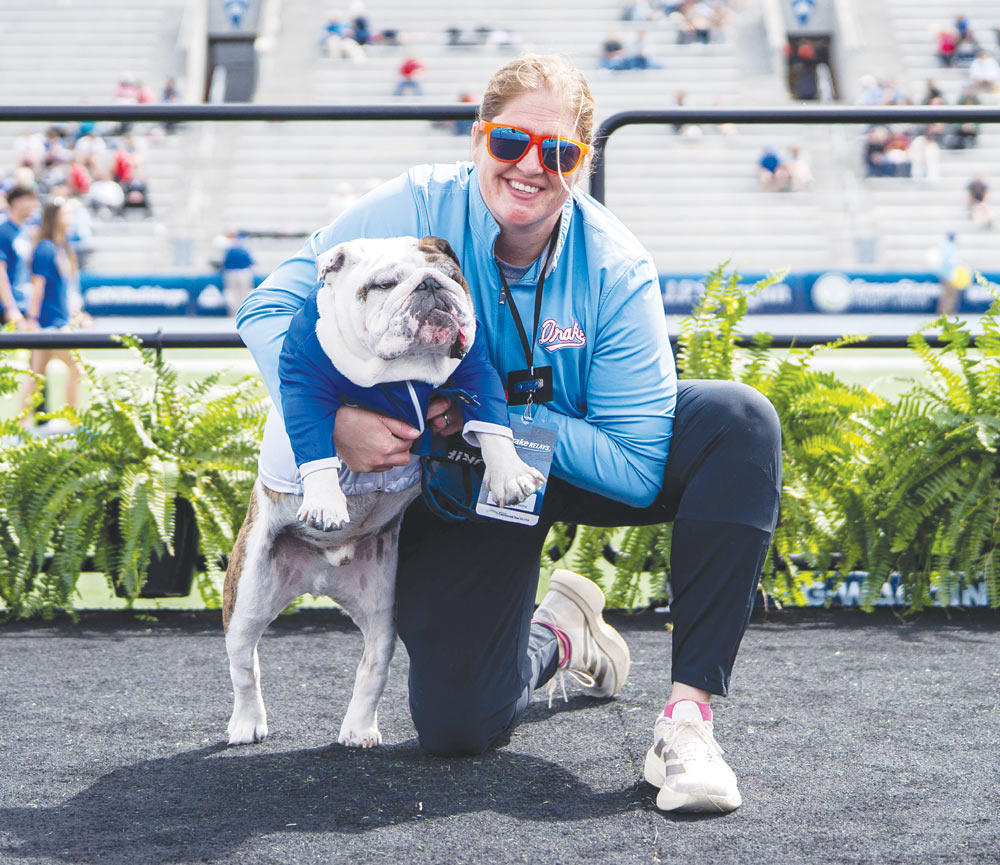 A smiling woman in sunglasses kneels down on a stadium track to hug an English bulldog.