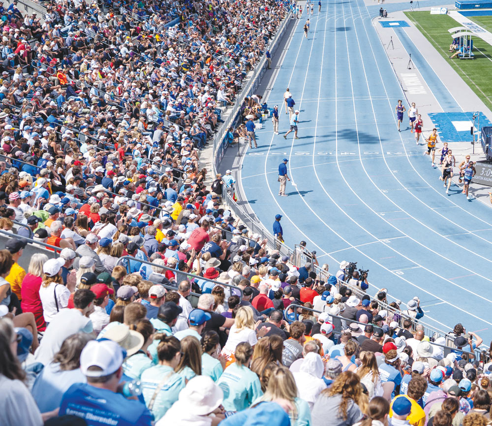 High-angle view of a packed stadium crowd watching runners compete on a blue track.