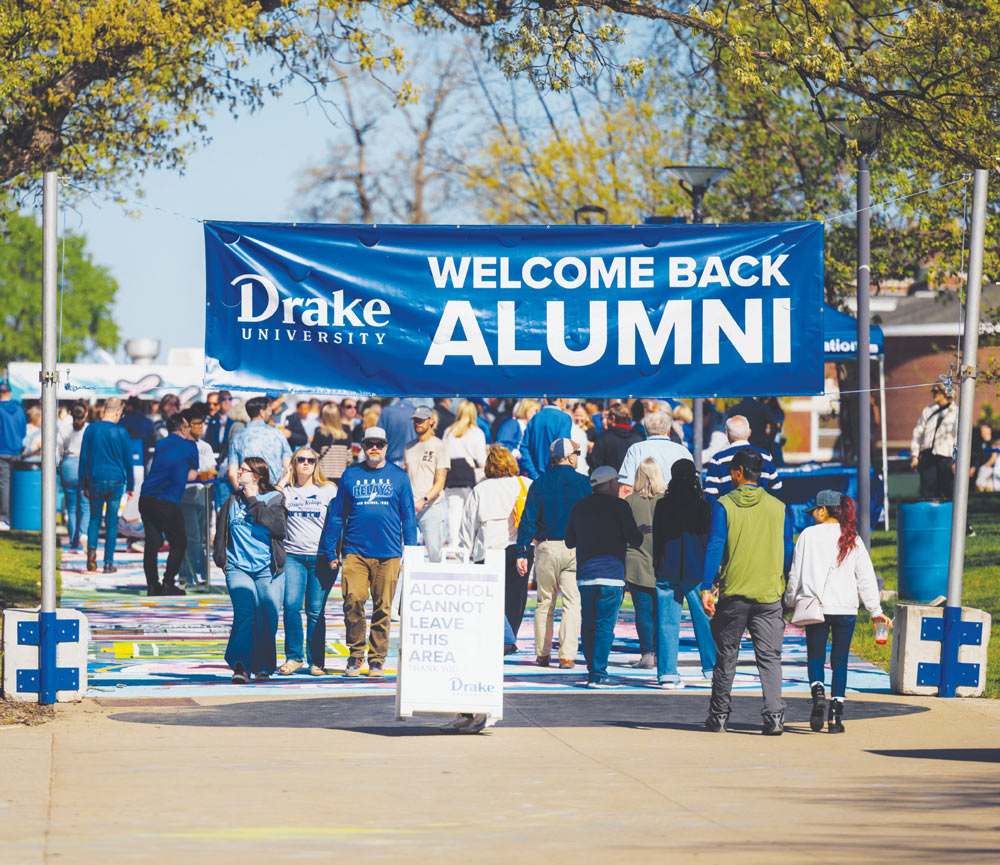 Crowd of people walking under a "WELCOME BACK ALUMNI" banner strung across a campus path.