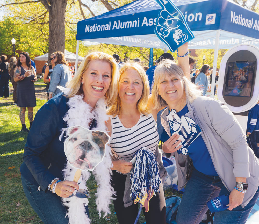 Three women pose with fun photo booth props, including a foam finger and bulldog cut-out, under an alumni tent.