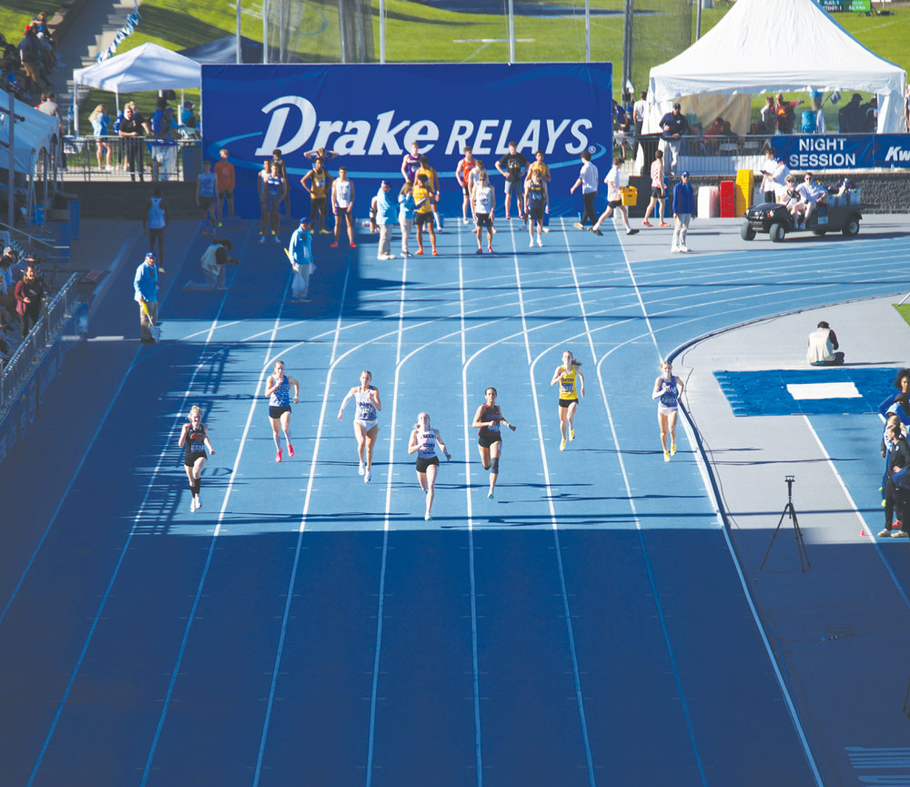 Sprinters race down a blue track during a competition under a large "Drake RELAYS" sign.