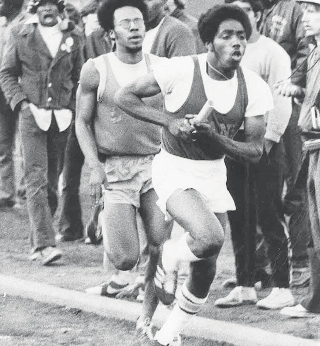 Black-and-white photograph of a runner carrying a relay baton, with another runner and spectators behind him.
