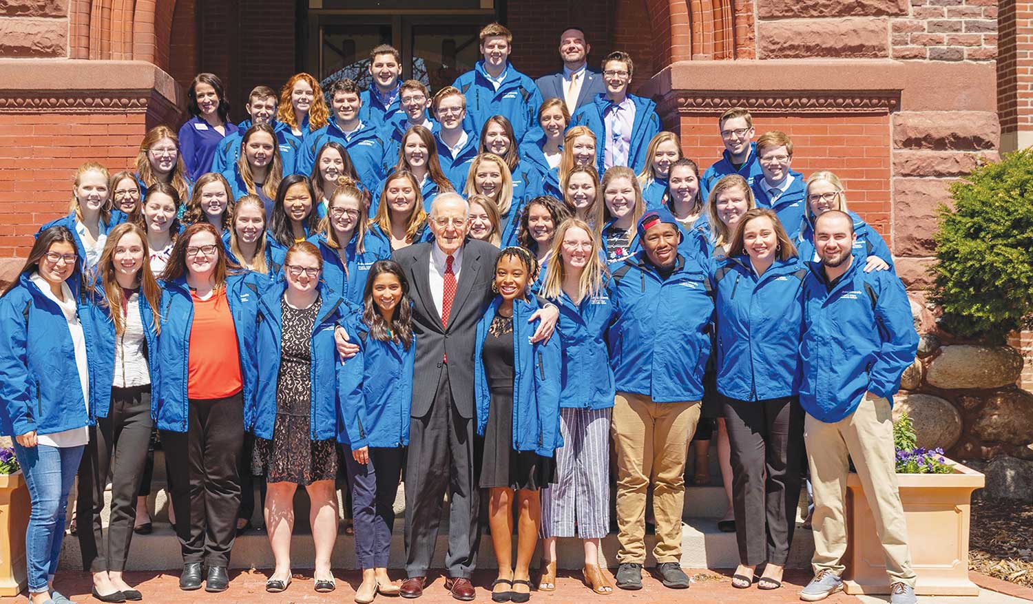 Large group of students in matching blue jackets gathered on steps with a gentleman in a suit.